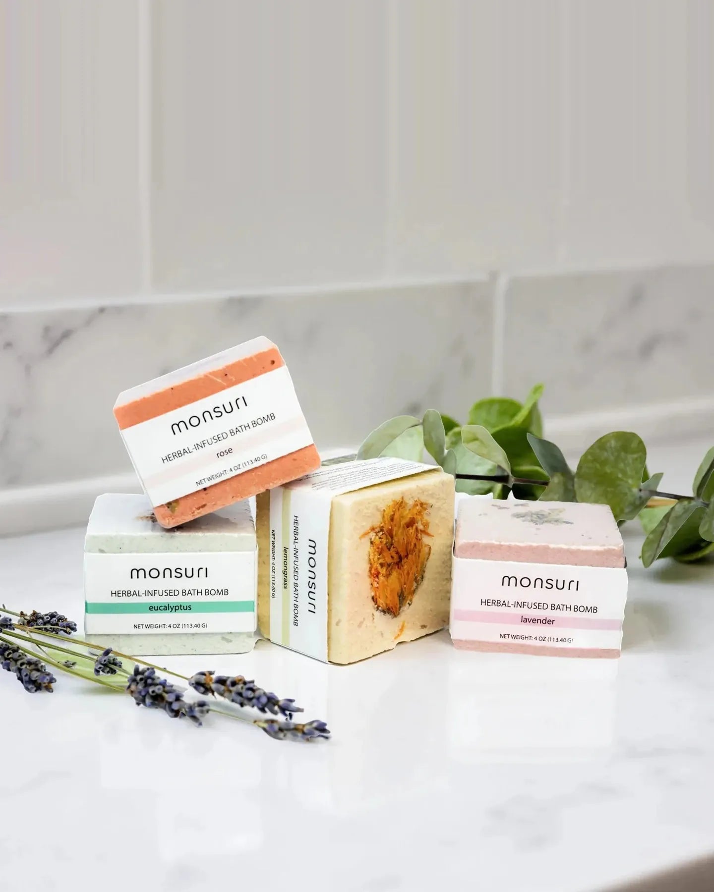 All four herbal-infused bath bombs on a marble counter with eucalyptus and dried lavender — rose, eucalyptus, lemongrass and lavender, each a 4 oz (113.40 g) cube in its own paper band, the lemongrass one turned to show the calendula pressed into its face.