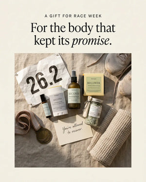 A gift for race week — the five recovery pieces laid on linen and styled with a 26.2 marathon bib, a finisher medal and a rolled waffle towel, beside a handwritten card reading "You're allowed to recover."
