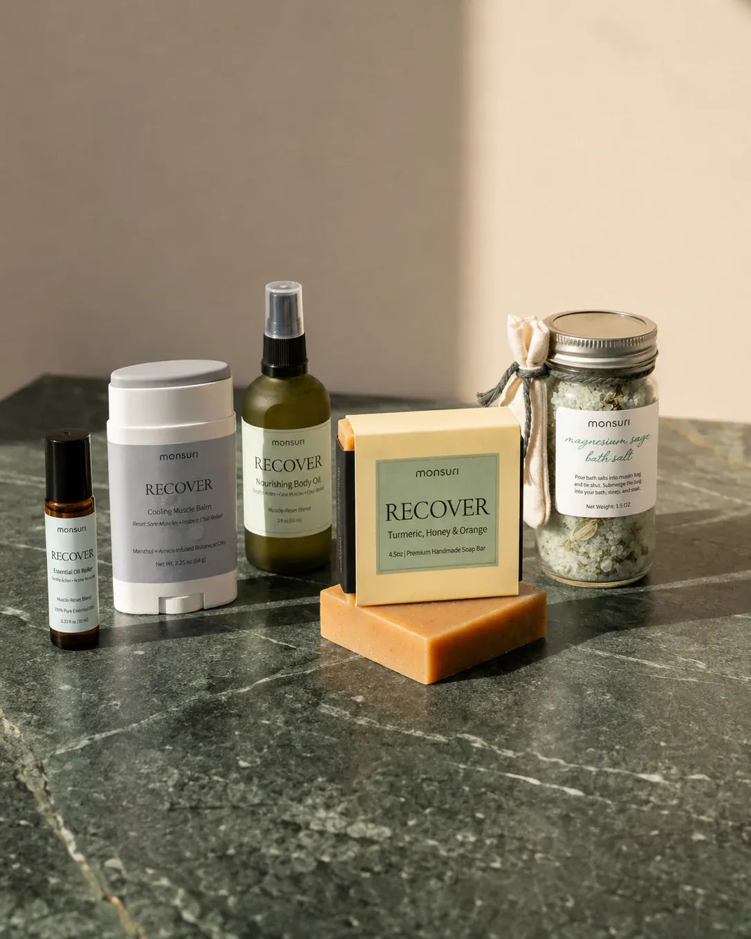 All five pieces of the post-workout recovery kit on a green marble counter — the 0.33 fl oz RECOVER essential-oil roller, the 2.25 oz cooling menthol and arnica balm stick, the 2 fl oz nourishing body oil in olive glass, the 4.5 oz turmeric, honey and orange soap bar, and the 1.5 oz magnesium sage bath salts in a lidded jar with its muslin bag.
