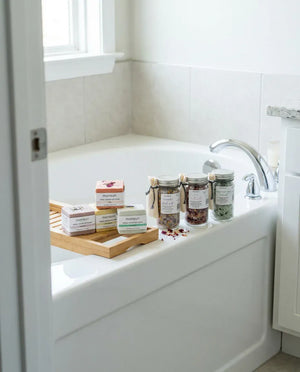 The bath bomb and salts set laid out along the edge of a bath in a bright bathroom, the four bath bombs and three salt jars arranged on a wooden caddy under a window.