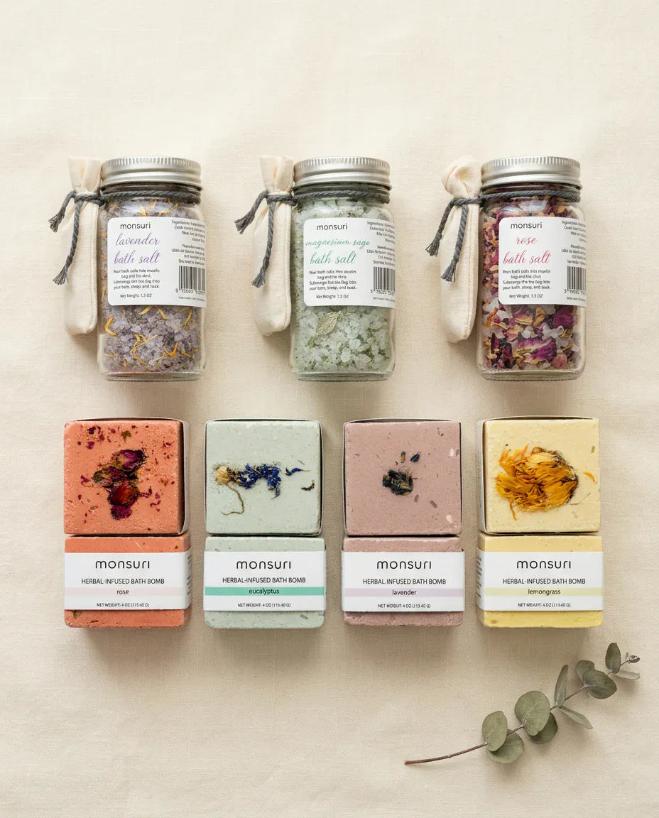 Flat lay of all seven pieces on cream linen: the lavender, magnesium and sage, and rose herbal bath salt jars in a row above the eucalyptus, lavender, lemongrass and rose bath bombs, with a dried eucalyptus stem alongside.