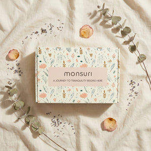 Overhead flat lay of the Monsuri botanical kraft gift box on rumpled cream linen, printed "A journey to tranquility begins here", with dried eucalyptus, lavender buds and dried rose petals scattered around it.