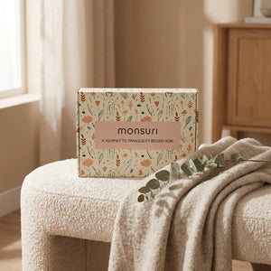 The Monsuri botanical kraft gift box resting on a cream boucle bench with an oatmeal wool throw and a dried eucalyptus stem, in soft window light.