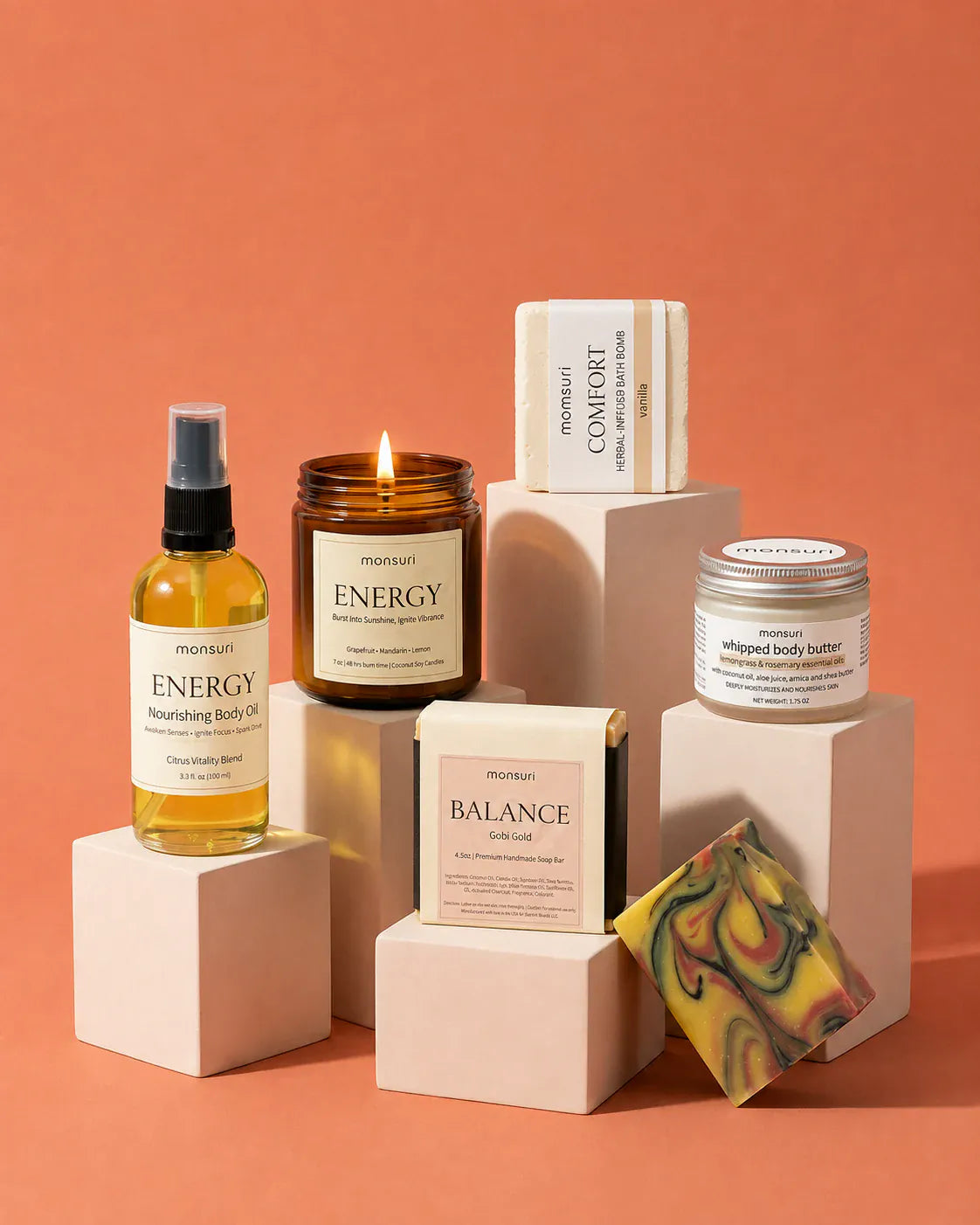 The five pieces staged on pale pink plinths against a terracotta backdrop, the ENERGY candle burning and the marbled soap bar propped at the front.