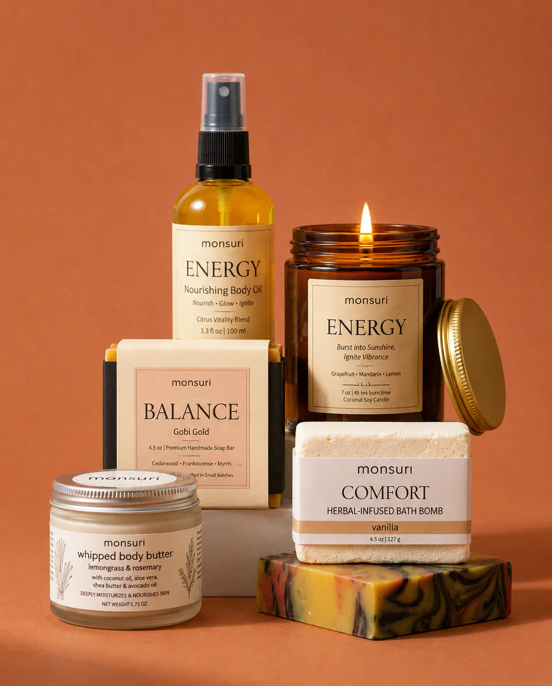 Close view against terracotta of the ENERGY nourishing body oil and the lit ENERGY grapefruit candle, with the whipped body butter, the boxed BALANCE soap and the COMFORT bath bomb behind them.
