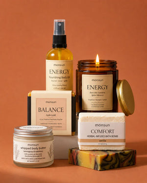 Close view against terracotta of the ENERGY nourishing body oil and the lit ENERGY grapefruit candle, with the whipped body butter, the boxed BALANCE soap and the COMFORT bath bomb behind them.