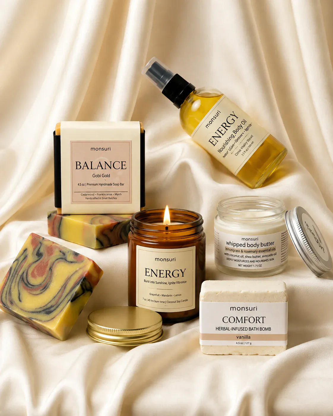The five pieces gathered on cream silk with the ENERGY candle lit, its gold lid set down beside the marbled Gobi Gold soap bar.