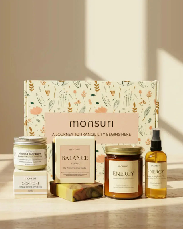 The Monsuri botanical kraft gift box with all five Bestie Boost pieces in front of it on a stone counter in a shaft of afternoon light — the 1.75 oz whipped body butter, the COMFORT vanilla bath bomb, the 4.5 oz BALANCE Gobi Gold soap with its marbled bar, the 7 oz ENERGY grapefruit candle and the 3.3 oz ENERGY nourishing body oil.