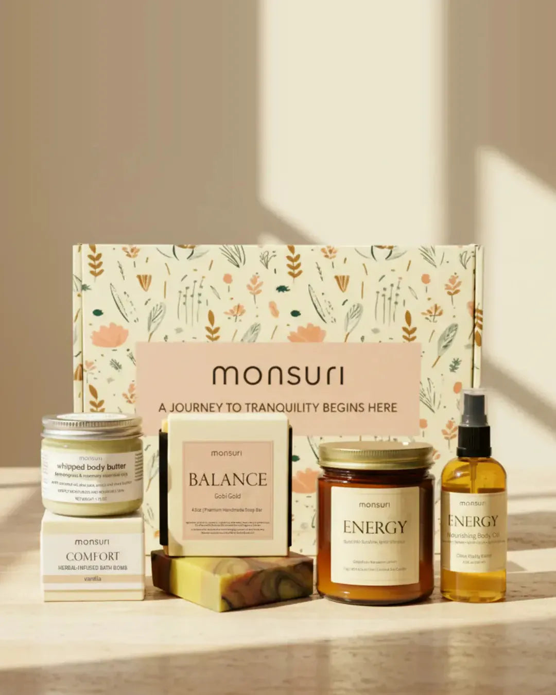 The Monsuri botanical kraft gift box with all five Bestie Boost pieces in front of it on a stone counter in a shaft of afternoon light — the 1.75 oz whipped body butter, the COMFORT vanilla bath bomb, the 4.5 oz BALANCE Gobi Gold soap with its marbled bar, the 7 oz ENERGY grapefruit candle and the 3.3 oz ENERGY nourishing body oil.
