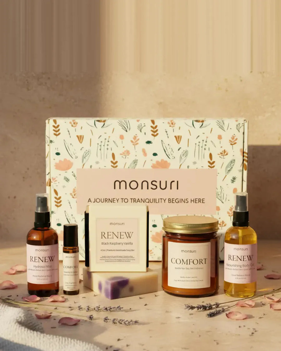 The five pieces of the Thinking of You gift set in front of the Monsuri botanical box on warm marble - the RENEW rose geranium hydrosol mist, the COMFORT essential oil roller, the 4.5 oz RENEW black raspberry vanilla soap, the 7 oz COMFORT vanilla amber candle and the RENEW nourishing body oil, with rose petals and lavender