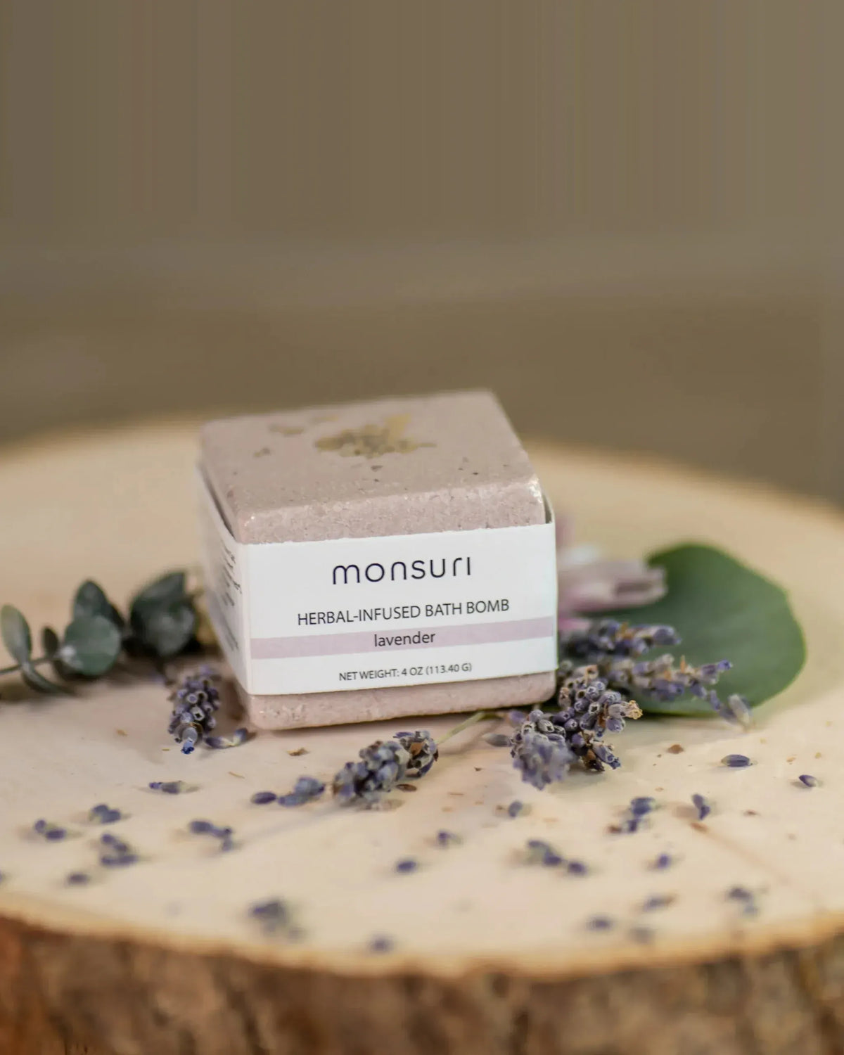 Close-up of the Monsuri lavender herbal-infused bath bomb, 4 oz, on a wood slice scattered with dried lavender buds and eucalyptus