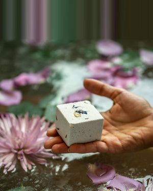 A hand holding a Monsuri herbal-infused bath bomb above a bath strewn with pink and purple petals, showing the size of the 4 oz bomb in the palm