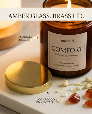 Infographic reading 'Amber Glass. Brass Lid.' highlighting the Monsuri COMFORT candle's light-protecting amber jar and brass lid beside jasmine blossoms.